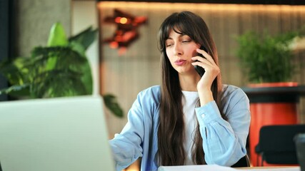 Professional young businesswoman wearing a light blue shirt, conversing on her smartphone while using a laptop in a modern office. She looks focused and cheerful. - Powered by Adobe