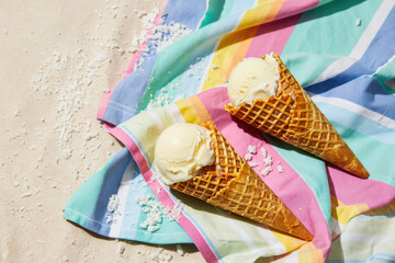 Two vanilla ice cream cones on a striped towel with coconut flakes on a sandy surface under bright light