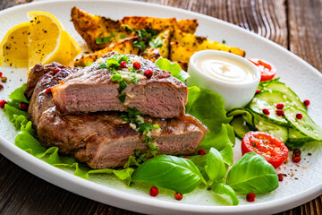 Grilled beef steak with baked potato wedges and fresh vegetables on wooden table	