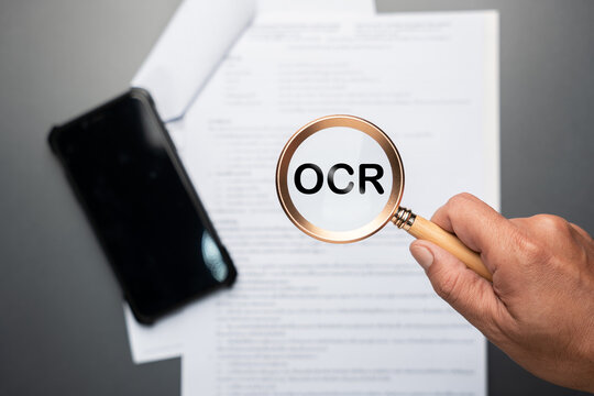 Hand holding magnifying glass with OCR text over document paper and smartphone. Symbolizes optical character recognition, text extraction, digitizing printed documents, and digital data conversion
