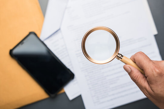 Hand holding magnifying glass over printed document with smartphone nearby. Concept of text scanning, document inspection, information analysis, and data extraction using digital tools