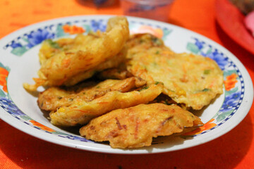 Plate filled with several pieces of fried fritters, a type of Indonesian or Southeast Asian street food, resting on a patterned tablecloth. Related to traditional snacks, comfort food, culinary themes