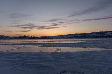 Fototapeta premium Serene winter landscape at dusk, showcasing frozen lake surface reflecting soft hues of orange and purple, surrounded by distant snow-capped mountains and tranquil atmosphere