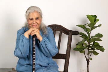 Indian retired senior woman with her wooden walking stick at home. Happy senior woman relaxing at home looking at camera. Smiling grandmother sitting on wooden chair at home