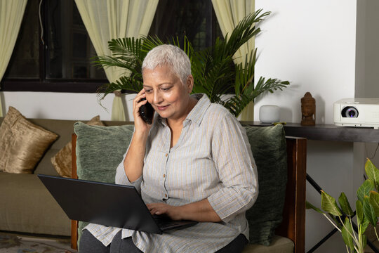Senior woman with cancer working on laptop 
while talking on mobile phone n her apartment.