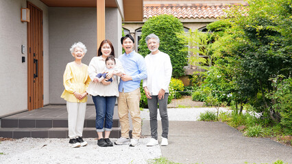 A three-generation family smiling and surrounding a baby in front of their house
