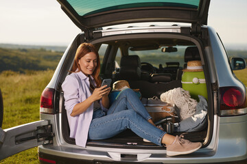 In a beautiful outdoor setting, a young woman relaxes on the tailgate of a car, enjoying a picnic...