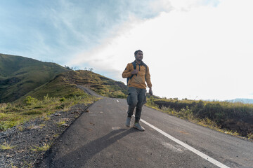 A man in outdoor gear walks confidently along a mountain road, holding a smartphone. The morning light and empty road highlight a sense of purpose, solitude, and discovery.