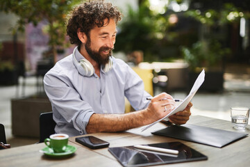 A man with headphones sits at a table in a trendy cafe, writing notes on paper while sipping...