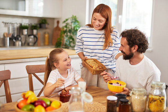 A joyful family shares breakfast in their kitchen, with the daughter smiling as she eats cereal, while the mother serves homemade bread to the father, creating a warm atmosphere.