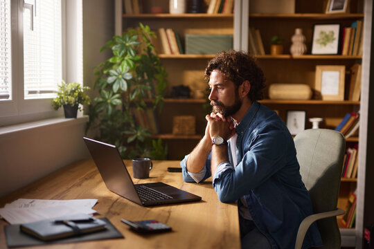 A man sits at a wooden desk in a sunlit home office, deeply engaged with his laptop. Surrounding him are plants and books, creating a warm, inviting workspace.