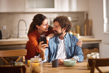 A couple smiles at each other while looking at a smartphone in their cozy kitchen. They are seated at a wooden table enjoying breakfast in a warm and inviting atmosphere.