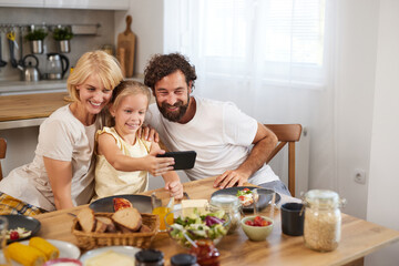 A cheerful family of three captures a selfie at their dining table filled with breakfast food in a...