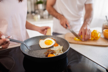 A couple engages in preparing a delightful breakfast. One person flips a fried egg, while the other slices fresh fruit on a wooden board in a bright kitchen.