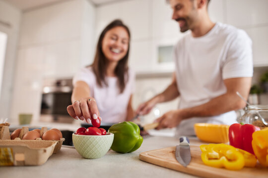 In a bright kitchen, a couple enjoys cooking together. One person reaches for cherry tomatoes while the other prepares ingredients, creating a joyful and collaborative atmosphere.
