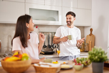 In a bright kitchen, a man holds a cup and smiles as he interacts with a woman. They are enjoying a casual breakfast surrounded by fresh fruits and a warm atmosphere.