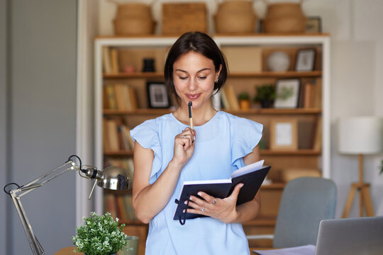 A woman stands in her home office, holding a notebook and pen, appearing thoughtful as she brainstorms ideas. The space is well-organized and inviting, with bookshelves in the background.