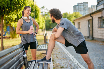 Two individuals are exercising in a park, with one stretching while the other smiles. They are...