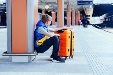 Teen boy sitting on platform with suitcase, observing arriving people