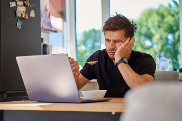 Man sitting at kitchen table, eats oatmeal porridge with displeased look and pushed her plate away. Unhappy man eating tasteless breakfast in kitchen table with laptop