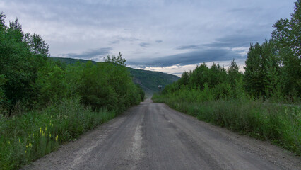 Fototapeta premium Dirt road stretches through lush greenery, flanked by dense foliage and distant hills under a cloudy sky, inviting exploration and adventure in a serene natural landscape