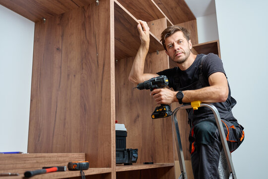 Man wearing work overalls assembling wooden wardrobe with drill and tools. Carpenter standing on step ladder, furniture installation process