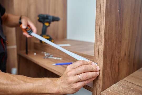 Close-up of man measuring wooden wardrobe with tape measure during furniture assembly, with tools and screws on shelf in background. Concept of home renovation and DIY furniture project