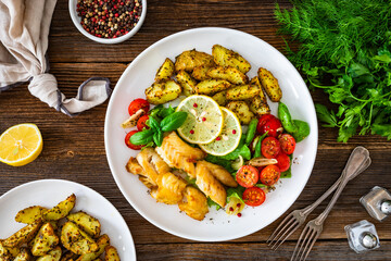Seared pollock fish, baked potato wedges and tomato salad on wooden table	