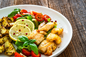 Seared pollock fish, baked potato wedges and tomato salad on wooden table	