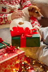Woman in festive sweater cuddles with small dog surrounded by wrapped Christmas gifts and glowing...