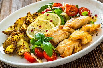 Seared pollock fish, baked potato wedges and tomato salad on wooden table	