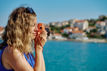 Woman standing near sea holding instant camera and taking photo. Female tourist at coastal resort...