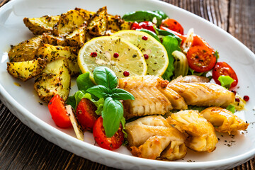 Seared pollock fish, baked potato wedges and tomato salad on wooden table	