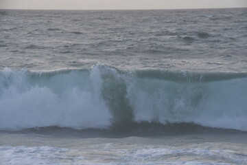 A powerful ocean wave is caught mid-break, with white foam forming at its crest. The sea is rough and textured, under a cloudy, grey sky, creating a dramatic and moody coastal scene