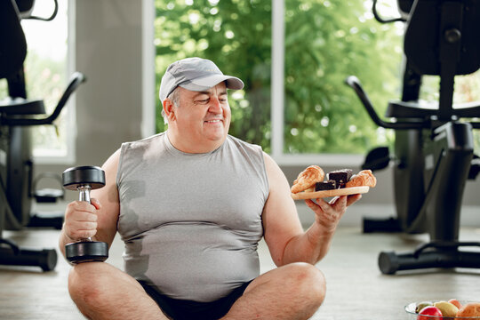 Man with dumbbell and Donut looking at camera. Happy fat guy holding free weights and eating big yummy hamburger. Sport, food, failed diet, workout exercise, cheat day concept