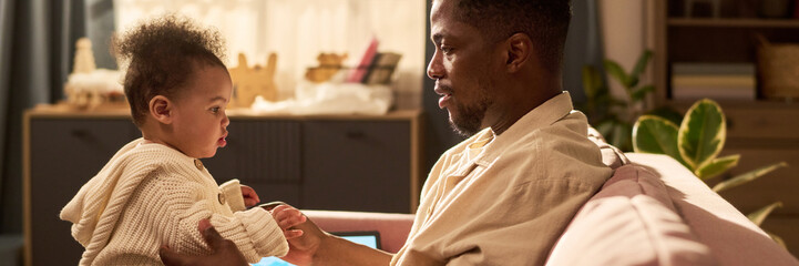 Black man sitting on sofa holding infant child facing him, engaging in bonding during paternity...