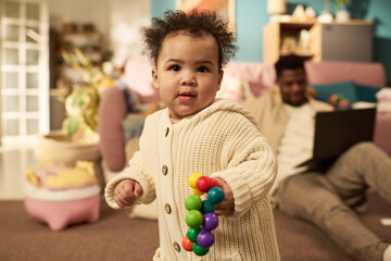 Portrait of baby playing with colorful toy in living room while Black man sitting on floor in background working on laptop, illustrating paternity leave and work life balance at home