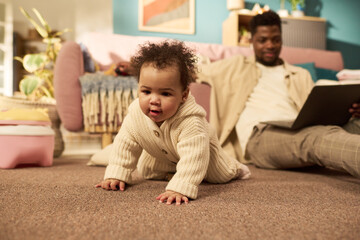 Infant crawling on carpet while Black young adult man sitting in background using laptop,...