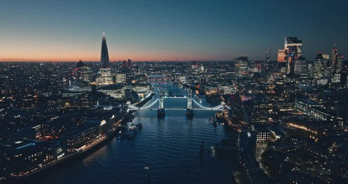 London: Aerial view of city illuminated skyline at sunset, iconic Tower Bridge, The Shard, and night cityscape stretching along the River Thames. Drone flight footage zoom out panorama. Blue toning