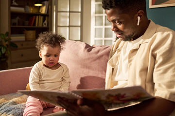 Black man spending paternity leave reading picture book to young child on sofa, both wearing casual...
