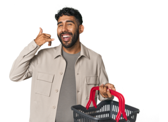 Hispanic male holding shopping basket in studio showing a mobile phone call gesture with fingers.
