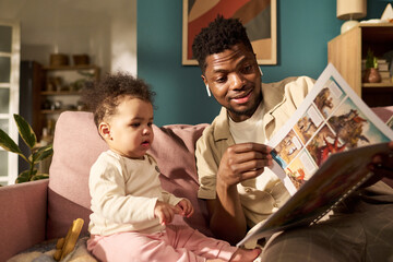 Black young adult man sitting on sofa reading picture book to toddler child during paternity leave,...