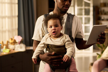 Black young adult man holding tablet while carrying baby in front carrier, standing indoors, focusing on multitasking and childcare during paternity leave, baby looking forward