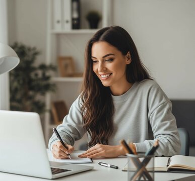 Happy young woman using laptop sitting at desk writing notes while watching webinar, studying online, looking at pc screen learning web classes or having virtual call meeting remote working from home.