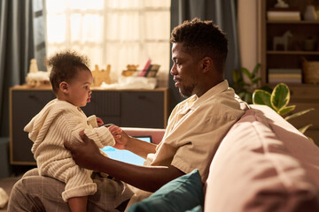 Black young adult man sitting on sofa holding and interacting with child in living room setting,...