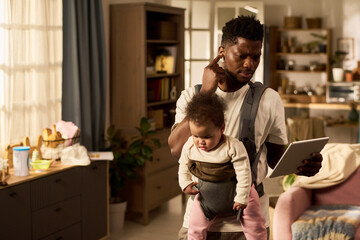 Black young adult man carrying infant child in baby carrier while multitasking with digital tablet in home setting, focusing on balancing paternity leave responsibilities and childcare
