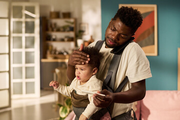Black young adult man multitasking with infant child in baby carrier, holding phone between ear and...