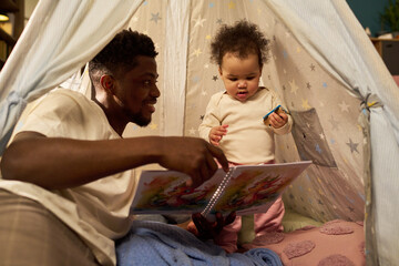 Black young adult man spending paternity leave reading storybook to baby inside play tent, both...