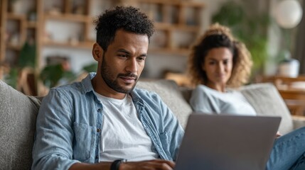On a couch a man uses a laptop while a woman is out of focus in the background