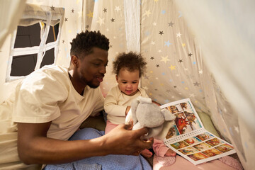 Black young adult man spending paternity leave reading picture book to baby child inside play tent, both sitting close together, father holding soft toy, child looking at book